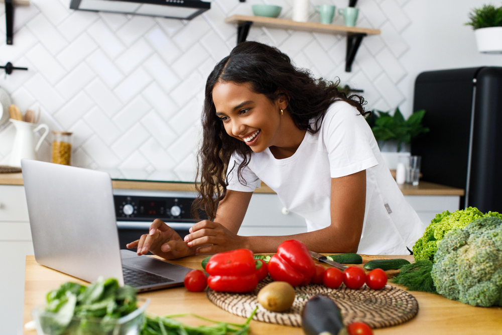 woman using laptop in kitchen surrounded by vegetables